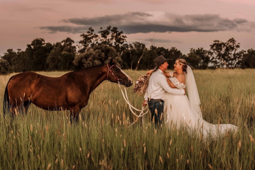 A man and a woman embrace on a dock with tall grass and a horse behind them leading the way